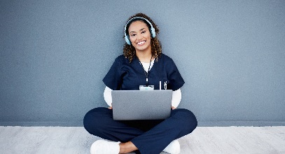 young lady with brown hair in blue scrubs sitting on the floor with a laptop in her lap