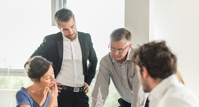 three men and one woman surrounding a table discussing what they are looking at