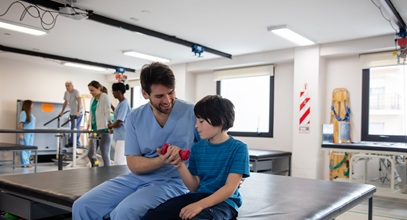 man in blue scrubs helping a small male child in a teal tshirt hold a weight while they are in a medical rehabilitation clinic