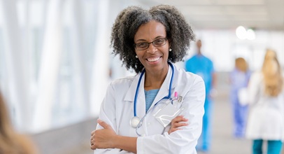 african american nurse in a white lab coat and stethoscope standing in a hospital hallway with her arms crossed smiling