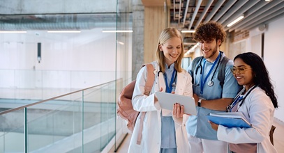 two young women, one blond and one dark haired, and one young man in a hallway wearing blue scrubs discussing paperwork