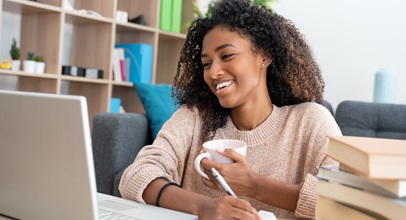 young african american woman drinking out of a mug, writing on a notepad while looking at her laptop with a pile of books stacked next to her