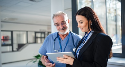 older white man in blue scrubs and younger woman with long brown hair discussing something on a tablet standing in a healthcare facility