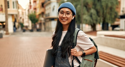 young lady with long dark hair carrying a backpack and wearing air pods holding a laptop standing in front of a building