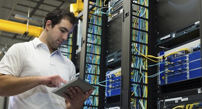dark haired man in a white polo looking at a tablet in a server room