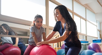 young woman in blue scrubs with long brown curly hair helping a young child balance on a pink balance ball