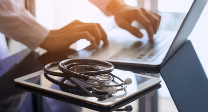 person typing on a laptop with a tablet and a stethoscope sitting next to them on the glass table