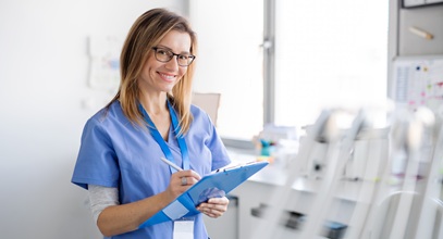 woman in blue scrubs and glasses writing on a clipboard in a hospital room
