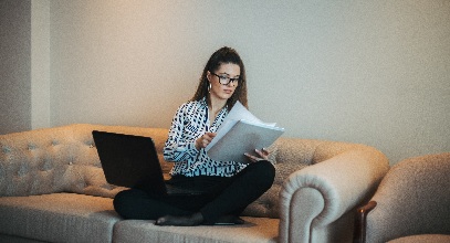 brown haired woman sitting crossed legged on a couch in a blouse and slacks with a laptop on her lap looking at papers