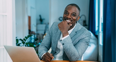 african american man in a grey jacket and white shirt smiling sitting at a desk with a laptop
