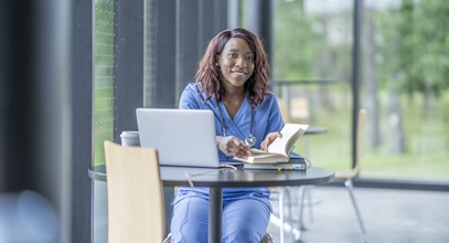 african american woman with long brown hair wearing blue scrubs is sitting at a table with an open book and a laptop