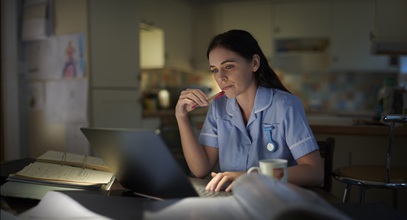 healthcare worker in blue scrubs and long brown hair looking at her laptop surrounded by open books
