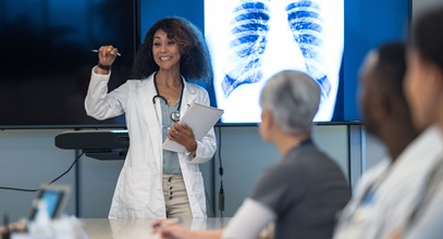 african american woman in a white doctor coat presenting to a room of other healthcare workers with an xray of a rib cage behind her
