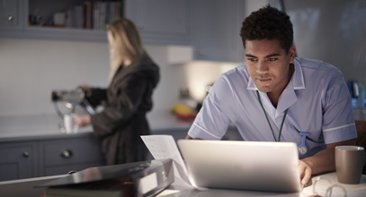 man in a blue shirt looking at a laptop with a blonde woman in a bathrobe in the background pouring coffee
