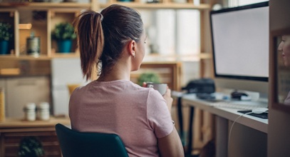 woman wearing a pink shirt and dark hair pulled back holding a mug sitting at a desk looking at a screen