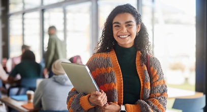 young woman with long brown curly hair and chunky orange sweater is holding a laptop standing in a classroom