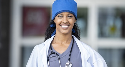 african american woman in scrubs and a white lab coat with a blue cap standing in a hallway