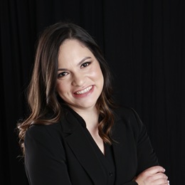 Laura Giraldo headshot in a black suit with arms crosses smiling at the camera