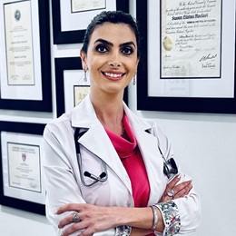 Roya Asghari standing in a hallway with multiple diplomas on the wall behind her, wearing a white coat and pink blouse